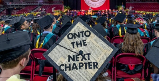 students at a commencement ceremony, with a graduation cap decorated to say Onto the Next Chapter in the center of the fram